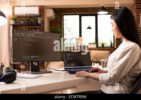 Focused female developer programming on laptop in home office. Asian woman building software and coding website remotely from modern apartment using digital tools for web development. Stock Photo