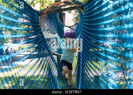 Cute child boy overcomes obstacles in rope tunnel outdoors. Modern amusement park for kids.  Stock Photo