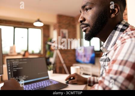 African American man sits at desk in modern apartment, concentrating on laptop and computer screens. Serious expression shows deep focus on code development and programming work. Stock Photo