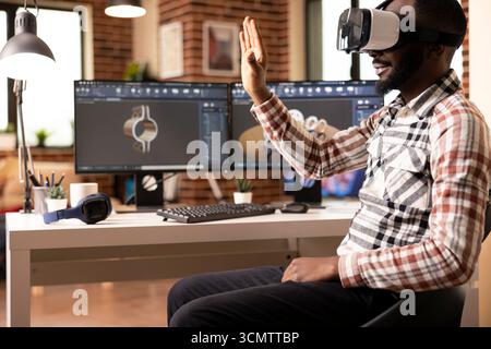 African american man wearing VR headset while seated at desk, remotely creating 3d clamps prototypes for construction company. Innovative design process using virtual reality tools. Stock Photo
