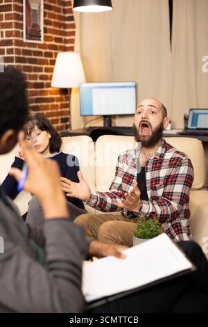 Caucasian man sits on sofa with wife, sharing marital challenges during therapy session. Black psychotherapist listens attentively while taking notes, supporting couple through relationship issues. Stock Photo