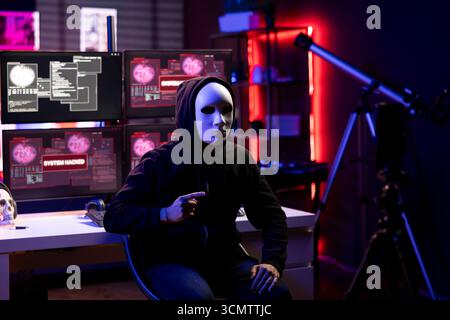 Masked man using phone to film video, doing espionage campaign from covert criminal den. Scammer wearing anonymous mask using smartphone to record demands from secret underground lair Stock Photo