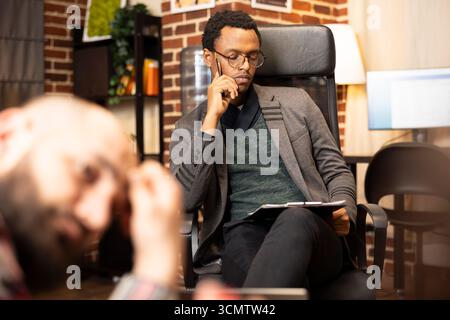 Thoughtful psychiatrist reads notes on clipboard during relationship assessment session. African american psychologist reviews notepad on knee and evaluates marriage struggles of couple in office. Stock Photo