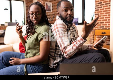 Tense black couple with cellphones seated on couch, not facing each other and expressing frustration through gestures. Boyfriend and girlfriend arguing in stylish brick wall home. Stock Photo