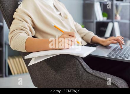 Sitting working in comfortable grey chair, focused work, writing in notebook typing on laptop Stock Photo