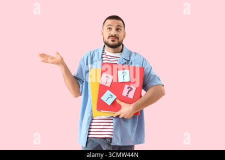 Portrait of thoughtful young man holding folders with question marks on pink background Stock Photo