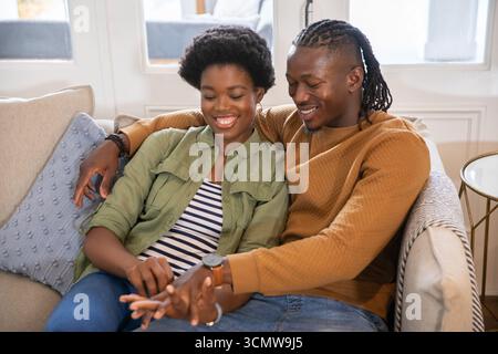 African american couple sitting on sofa in living room exchanging ring beside blue cushion Stock Photo