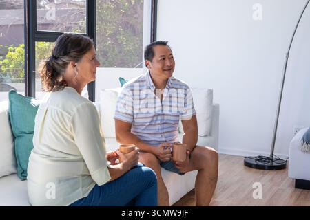 Diverse couple sitting on sofa by window in living room holding coffee mugs Stock Photo