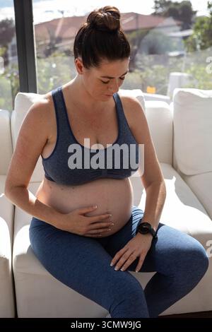 Pregnant woman sitting on white sofa by glass window wearing blue sportswear showing fitness watch Stock Photo