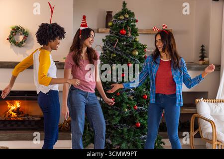 Diverse female friends gathering around christmas tree with ornaments by lit fireplace Stock Photo