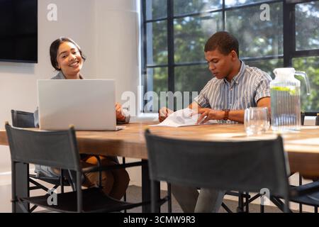 Diverse coworkers smiling and reviewing documents at office with laptop, lemon water pitcher Stock Photo