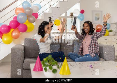 Diverse mom and teenage daughter sitting on sofa at home, popping black confetti poppers Stock Photo