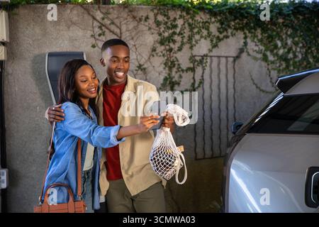 African American couple taking selfie with smartphone at carport holding grocery bag by charger Stock Photo