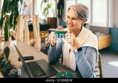 Happy senior woman with gray hair enjoying coffee at cafe while working remotely on laptop Stock Photo