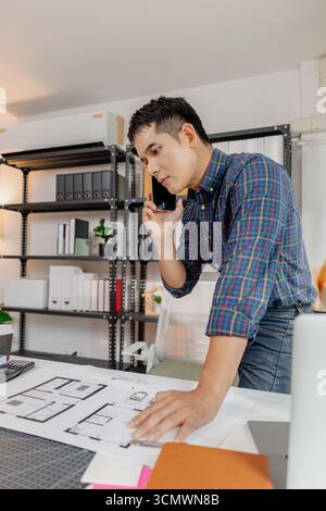 Confident young architect working at office desk, drawing house plans and discussing construction details on the phone, focusing on accuracy and creat Stock Photo