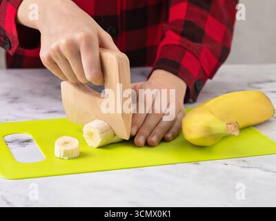 Child cutting a banana with a safe wooden knife on a plastic cutting board in green Stock Photo