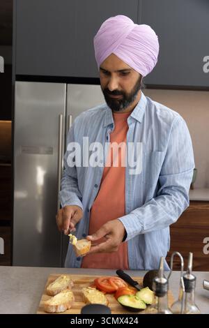Indian man spreading butter on bread on wooden cutting board at kitchen island with avocado halves Stock Photo