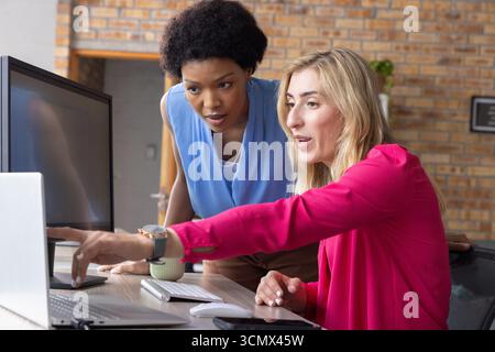 Diverse female coworkers collaborating at office desk with laptop and external monitor Stock Photo