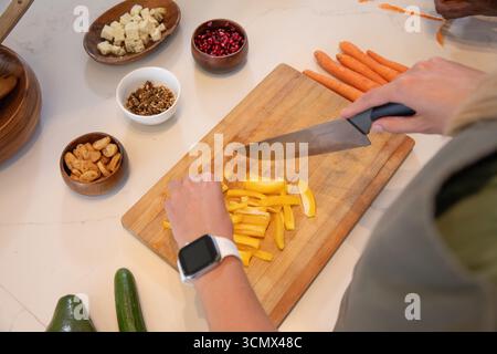 Friends slicing yellow pepper with chef knife on cutting board beside carrots in kitchen Stock Photo