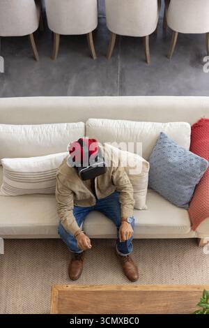 Indian man using vr headset on cream sofa with cushions near coffee table, potted plant Stock Photo