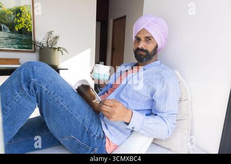Mid adult indian man wearing turban reclining on lounge chair at window holding magazine, mug Stock Photo