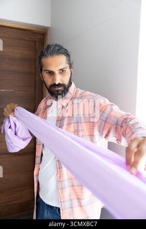 Mid adult indian man holding, stretching light purple fabric in room wearing pink plaid shirt Stock Photo