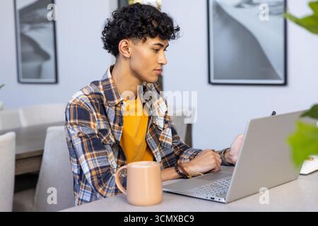 Man sitting at home desk working on laptop with stylus, coffee mug, prints and plant Stock Photo