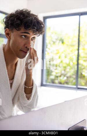 Man standing at bathroom wearing bathrobe wiping cheek with cotton pad by window, copy space Stock Photo