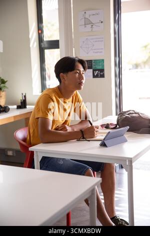 Asian teenage male student writing in notebook at classroom desk using tablet under natural light Stock Photo