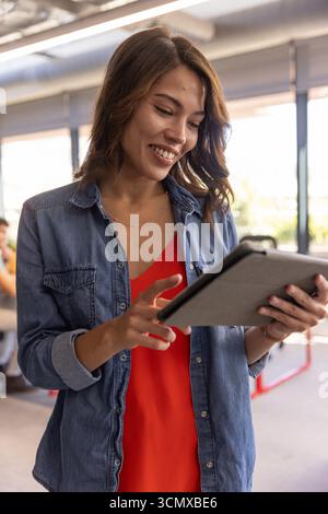 Hispanic woman tapping tablet screen while standing in office with blinds Stock Photo