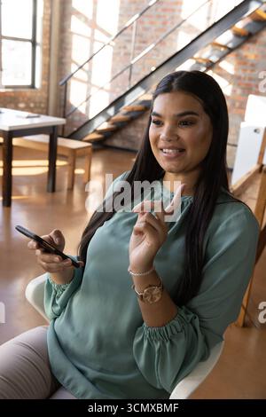Woman in her thirties gesturing while holding smartphone on white armchair in office loft Stock Photo