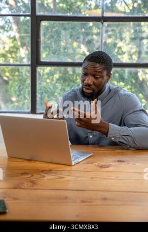 African american man leaning, gesturing at laptop on wooden table in office by framed window Stock Photo