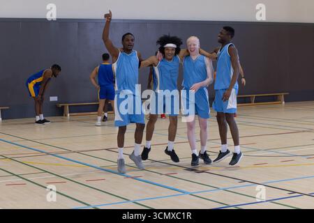 Diverse male basketball teammates in uniforms cheering and shouting on gym court with lines Stock Photo