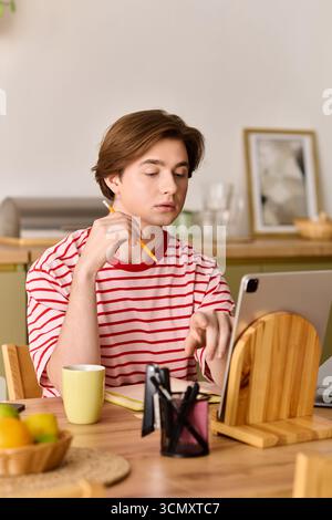 A young man studies intently at home, using a tablet while sitting at a wooden table. Stock Photo