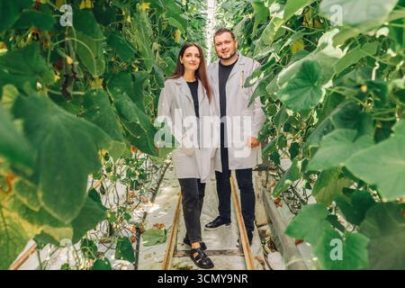Two individuals in gray lab coats standing among lush green plants in a greenhouse, showcasing teamwork and collaboration in agricultural innovation Stock Photo