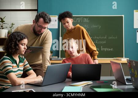 Caucasian man assisting diverse group of teenagers working on laptops in classroom, students focusing on screens while teacher holding digital tablet and explaining task Stock Photo