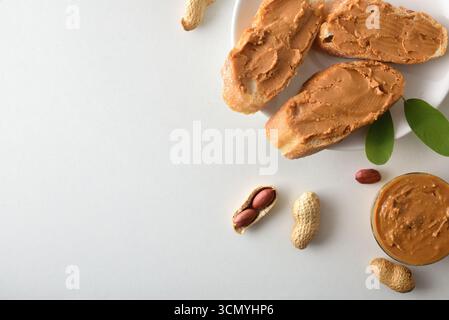 Detail of slices of bread with peanut butter on a plate and bowls of peanuts on a white table. Top view. Stock Photo