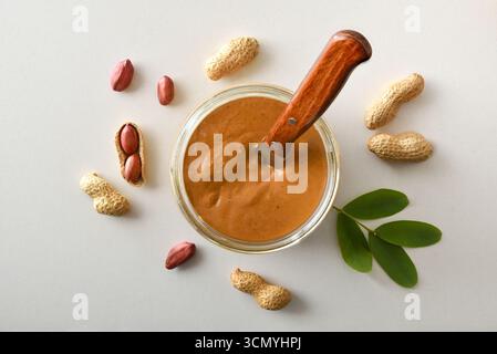 Detail of peanut butter in a glass jar with a metal and wooden spatula, with shelled peanuts and opened peanuts showing kernels with leaves for decora Stock Photo