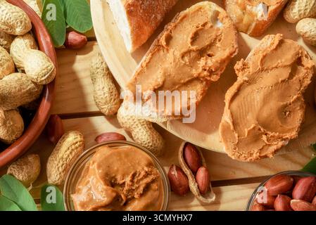 Detail of bowl of freshly harvested homemade peanut butter and slices of bread on a wooden table with fruit outside in the sun. Top view. Stock Photo