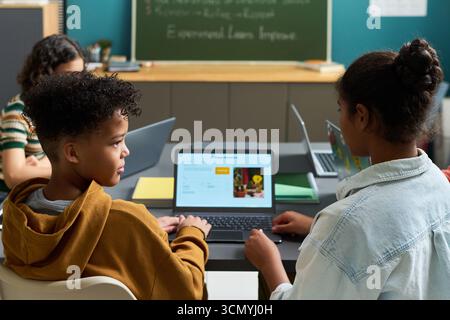 Black boy and Black girl sitting at desk using laptop computers in classroom, engaging in collaborative learning activity with other students visible in background Stock Photo