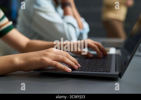 Teenager girl typing on laptop keyboard in classroom setting, focusing on screen while another student sits nearby, hands and arms visible, engaged in academic activity Stock Photo