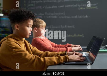 Multiethnic boys sitting side by side using laptops in classroom, focusing on typing and coding, with chalkboard and programming code visible in background, representing technology education Stock Photo