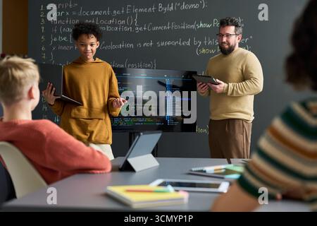 Teenager boy presenting programming project using tablet while standing near Caucasian male teacher holding digital tablet in classroom with students listening Stock Photo