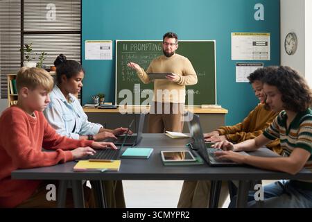 Caucasian man teaching diverse group of teenagers using laptops in classroom, students focusing on typing while instructor explaining lesson at chalkboard, educational technology concept Stock Photo