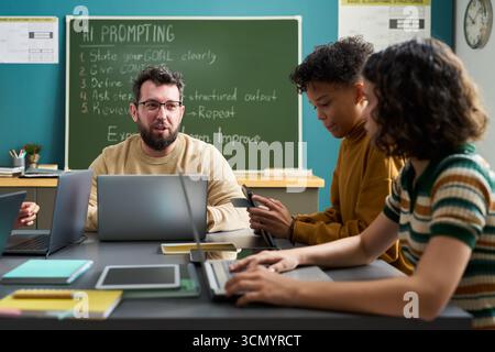 Caucasian young adult man explaining AI prompting to two multiethnic teenagers using laptops in classroom, students listening attentively and taking notes with digital devices Stock Photo