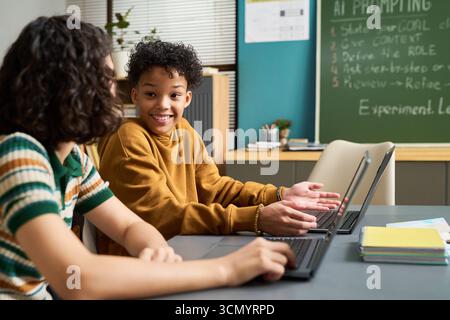 Two teenagers, one Black boy and one Caucasian girl, sitting at table using laptops, smiling and gesturing while collaborating on project in classroom with chalkboard in background Stock Photo