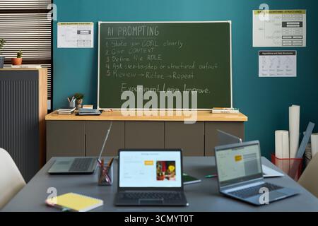 Classroom scene showing empty desks with open laptops in foreground, green chalkboard with AI prompting instructions written in chalk, educational posters on blue wall in background Stock Photo