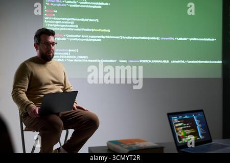 Caucasian man sitting on stool using laptop while presenting programming code projected on wall, holding computer on lap, explaining software development concepts Stock Photo