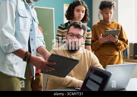 Caucasian man sitting at desk assisting multiethnic teenagers using digital tablets in classroom setting, students standing around teacher engaging with technology Stock Photo