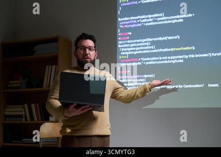 Caucasian young adult man presenting programming code on projection screen while holding laptop, standing in office setting, explaining software development concepts to audience Stock Photo